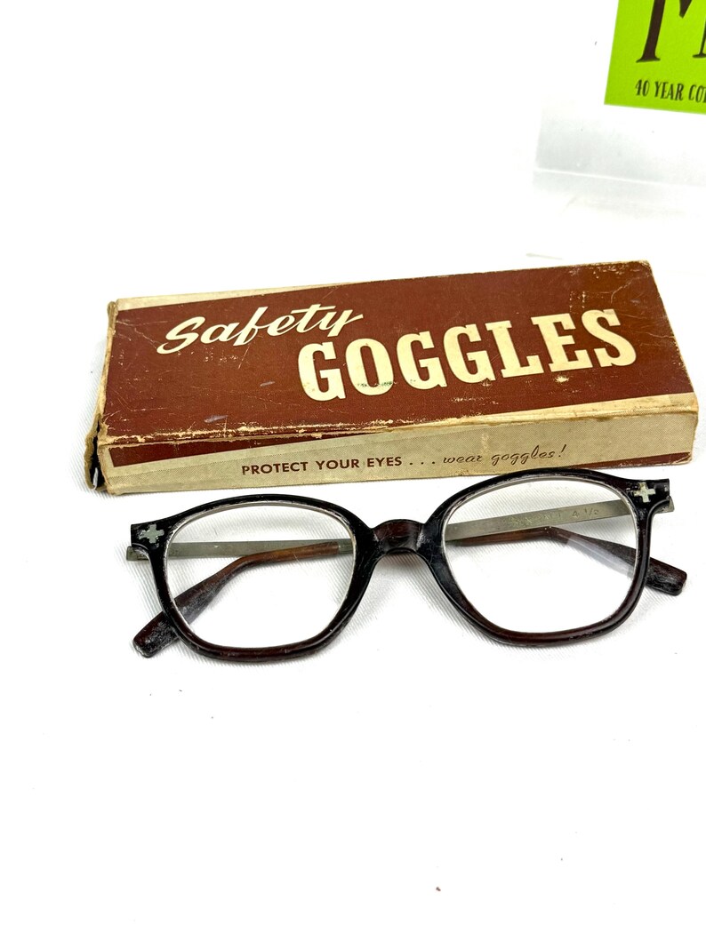 Pode incluir: &Oacute;culos de seguran&ccedil;a vintage com uma caixa castanha. A caixa diz "Safety Goggles" em letras brancas. Os &oacute;culos s&atilde;o pretos com lentes transparentes e um desenho de cruz nos cantos. O texto "PROTECT YOUR EYES... wear goggles!" est&aacute; na caixa.