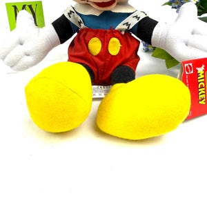 May include: A plush Mickey Mouse doll with a cream-colored face, black ears, and white gloves. He wears a blue shirt, red shorts with yellow buttons, and yellow shoes. A red tag with the word "Mickey" is visible.