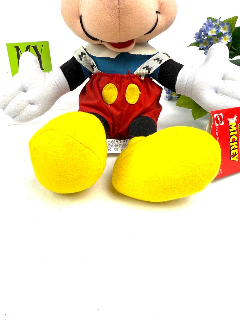 May include: A plush Mickey Mouse doll with a cream-colored face, black ears, and white gloves. He wears a blue shirt, red shorts with yellow buttons, and yellow shoes. A red tag with the word "Mickey" is visible.