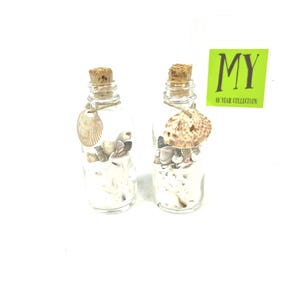 May include: Two small glass bottles with cork stoppers filled with seashells and sand. The bottles are tied with twine and have a seashell charm attached. The bottles are on a white background. The text "MY 10 YEAR COLLECTION" is on a green label.
