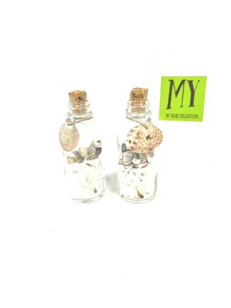 May include: Two small glass bottles with cork stoppers filled with seashells and sand. The bottles are tied with twine and have a seashell charm attached. The bottles are on a white background. The text "MY 10 YEAR COLLECTION" is on a green label.