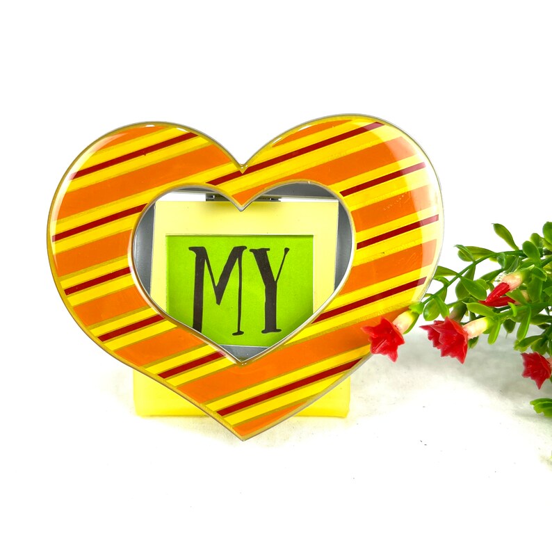 May include: A heart-shaped picture frame with orange and yellow stripes. The frame has a clear opening for a photo and the word "MY" is written on a green paper inside the frame.