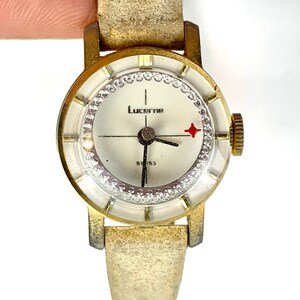 May include: Vintage Lucerne watch with a gold-tone case and a cream-colored face. The watch features a crystal-studded bezel, a red star accent, and a gold-colored band. The watch face displays the word "Lucerne" and "Swiss".