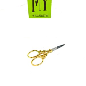May include: Gold-colored small scissors with ornate handles and a silver blade. The scissors are on a white background.  The text "MY 40 YEAR COLLECTION" is on a green square.