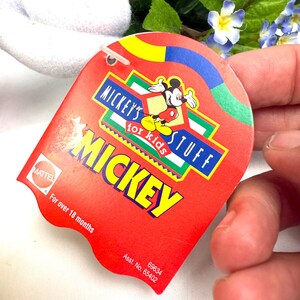 May include: A red tag with the text "MICKEY'S STUFF for kids MICKEY" and a graphic of Mickey Mouse. The tag also includes the text "For over 18 months" and the Mattel logo.