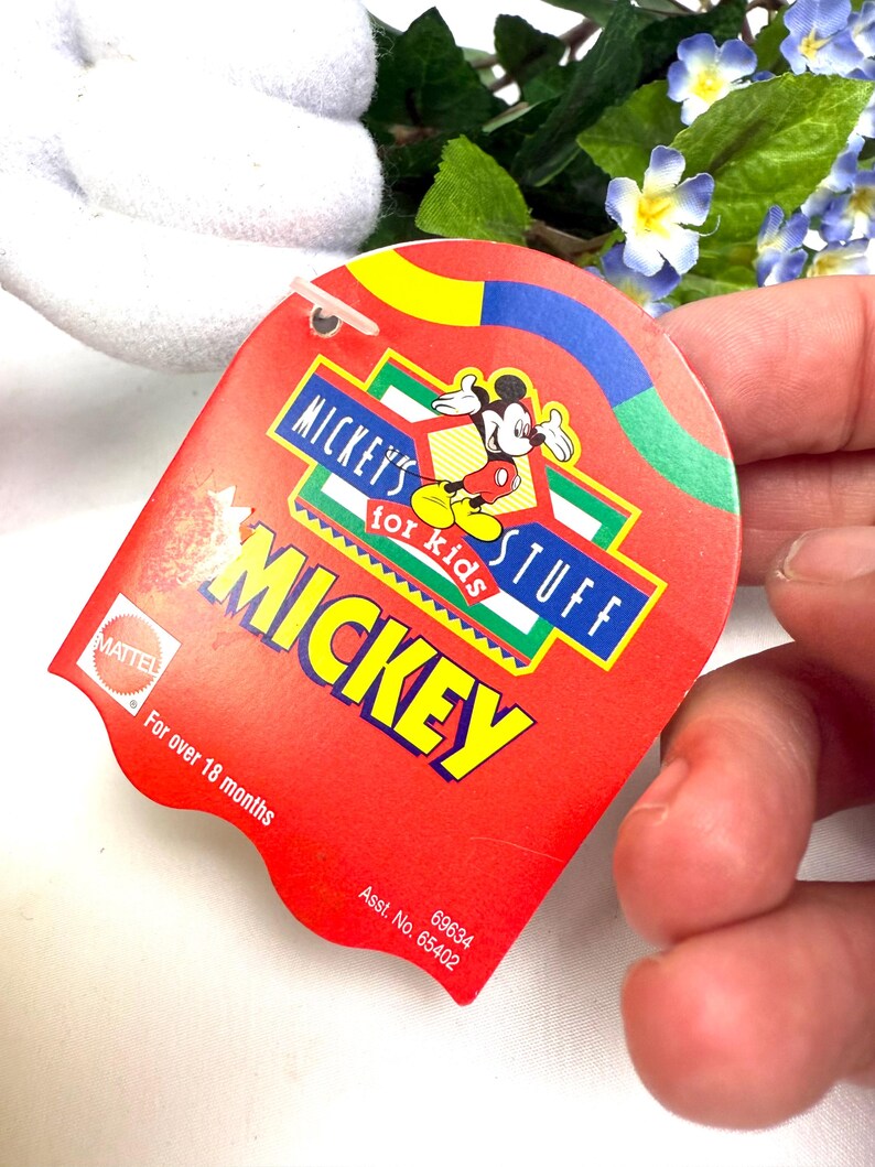 May include: A red tag with the text "MICKEY'S STUFF for kids MICKEY" and a graphic of Mickey Mouse. The tag also includes the text "For over 18 months" and the Mattel logo.