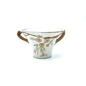 May include: White ceramic planter with two brown handles and floral relief details. The planter features a delicate design of pink flowers and green leaves, adding a touch of nature-inspired elegance. The handles are textured, providing a rustic contrast to the smooth white surface.