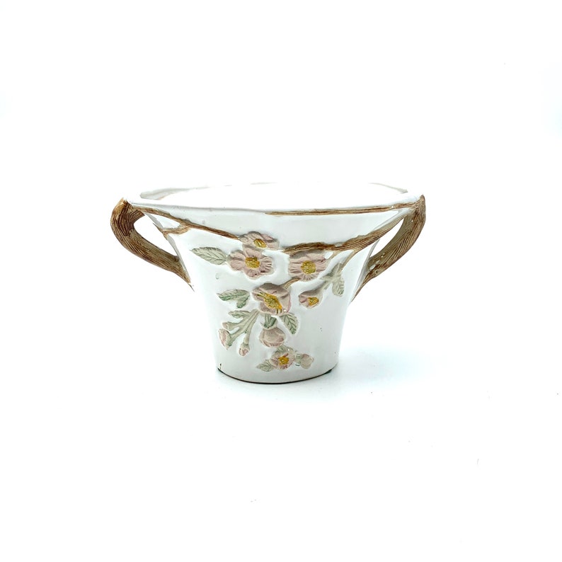 May include: White ceramic planter with two brown handles and floral relief details. The planter features a delicate design of pink flowers and green leaves, adding a touch of nature-inspired elegance. The handles are textured, providing a rustic contrast to the smooth white surface.