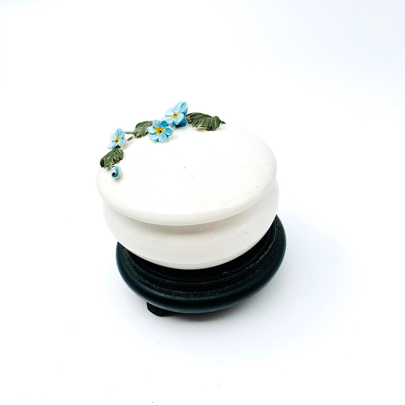 May include: A white ceramic trinket box with a floral design. The box has a lid and sits on a black wooden base.