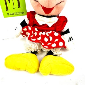 May include: A Minnie Mouse plush doll wearing a red dress with white polka dots, a white collar, and yellow shoes. The doll has a smiling face with a red bow and black eyes. A green tag with the text "MT 40 YEAR COLLECTION" is visible.