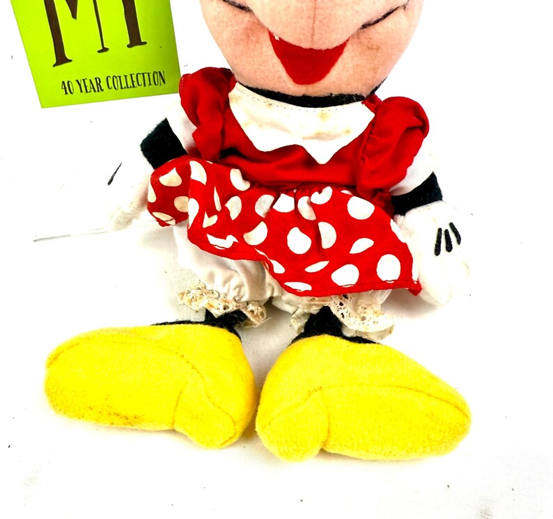 May include: A Minnie Mouse plush doll wearing a red dress with white polka dots, a white collar, and yellow shoes. The doll has a smiling face with a red bow and black eyes. A green tag with the text "MT 40 YEAR COLLECTION" is visible.