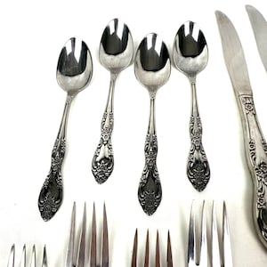 May include: An assortment of vintage silverware, including spoons, forks, and knives, arranged on a white surface. The silverware has ornate floral detailing on the handles, with a shiny, reflective finish. The spoons are oval-shaped.