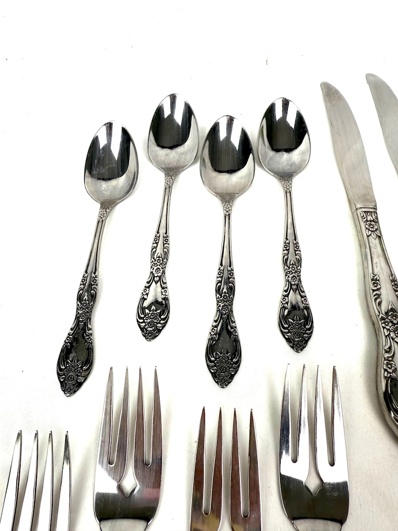 May include: An assortment of vintage silverware, including spoons, forks, and knives, arranged on a white surface. The silverware has ornate floral detailing on the handles, with a shiny, reflective finish. The spoons are oval-shaped.