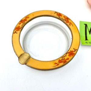 May include: A round glass ashtray with a gold rim and a floral design. The design features orange flowers and branches on a cream background.