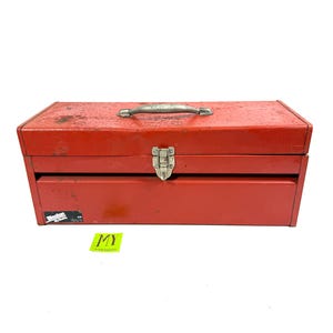 May include: A vintage red metal toolbox with a silver handle and clasp. The toolbox has a rectangular shape with a hinged lid and a lower drawer. The metal shows signs of wear and tear, with some rust and scratches visible. A small sticker is on the side.