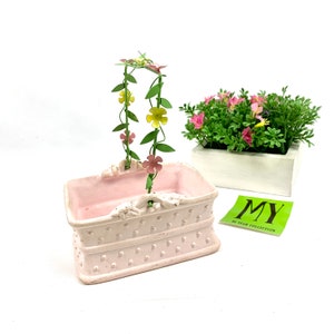 May include: A pink ceramic planter with a decorative design and a metal floral arch with pink and yellow flowers. The planter is sitting on a white surface.  MY 40 YEAR COLLECTION.