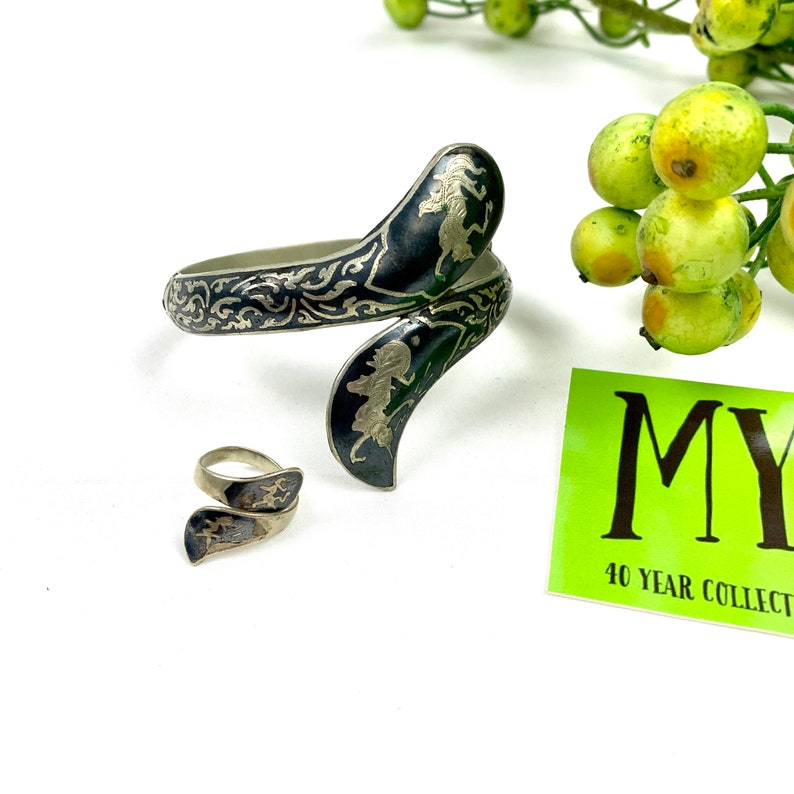 May include: A silver bracelet and ring set with a black enamel finish and intricate floral designs. The bracelet is a curved, open design and the ring is a simple band with a similar design.
