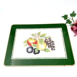 Op de afbeelding: Groene placemat met een gouden rand met een geschilderd ontwerp van fruit, waaronder een peer, een appel en pruimen. Het fruit wordt omringd door groene bladeren en witte bloemen.