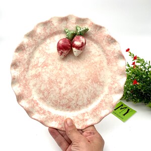 May include: A pink ceramic plate with a scalloped edge and a marbled design. Two small ceramic radishes are attached to the top of the plate.