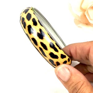 May include: A bangle bracelet featuring a glossy finish with a yellow and black leopard print pattern. The bracelet has a smooth, rounded exterior surface and a metallic interior lining, showcasing a classic circular design for the wrist.