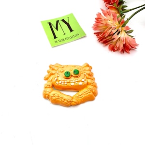 Pode incluir: Um broche extravagante em forma de caranguejo laranja com olhos verdes e dentes brancos. O broche está sobre uma superfície branca, com uma etiqueta verde que diz "MY 40 YEAR COLLECTION". Flores cor-de-rosa no fundo.