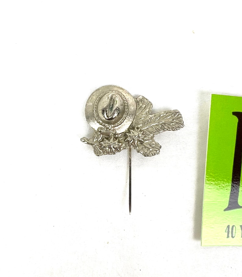 May include: Silver-toned pin featuring a cowboy hat with a feather design. The pin is attached to a thin, silver-toned pin.