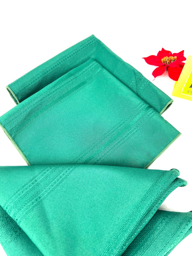May include: Three green linen napkins with a subtle striped pattern. The napkins are folded and stacked on top of each other.