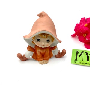 May include: A small ceramic figurine of a girl elf wearing a pink hat and orange dress. The elf has blonde hair and large eyes.