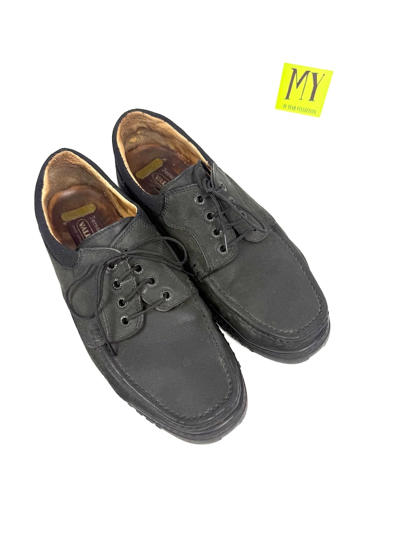 May include: A pair of black leather shoes with a lace-up design. The shoes have a rounded toe and a thick sole. The interior of the shoes is a light brown color. The shoes are displayed on a white background. A yellow sticker with the letters "MY" is in the upper right corner.