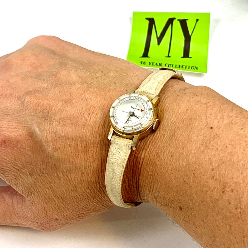 May include: A gold-toned Lucerne watch with a white face and a cream-colored leather band. The watch face has a star emblem and a second hand. The watch is on a wrist.