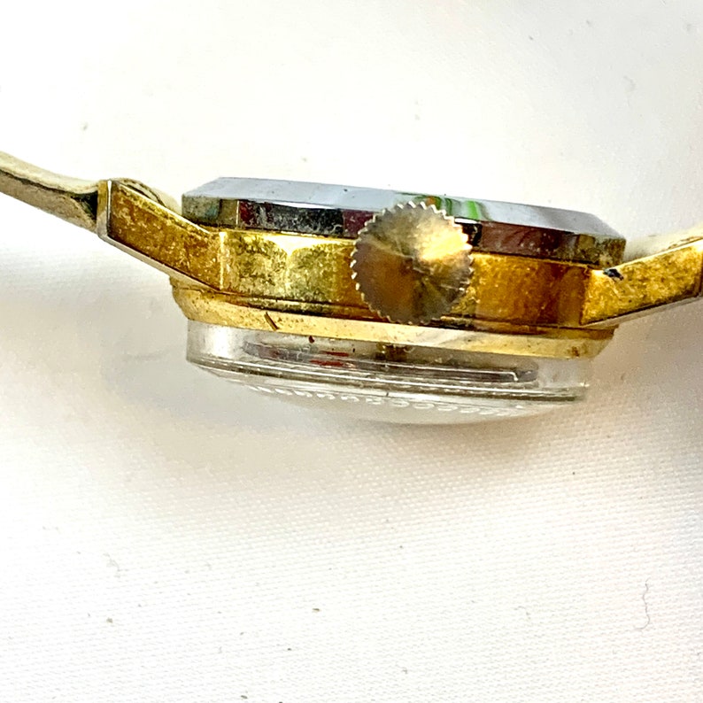 May include: Side view of a vintage gold-toned wristwatch with a silver-colored face and a clear, domed crystal. The watch has a small, textured crown and a thin, light-colored band.