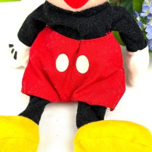May include: A plush Mickey Mouse doll with a black body, red shorts with white buttons, yellow shoes, and white gloves. The doll has a smiling face with a red mouth and black eyes. The doll is a classic Disney character.