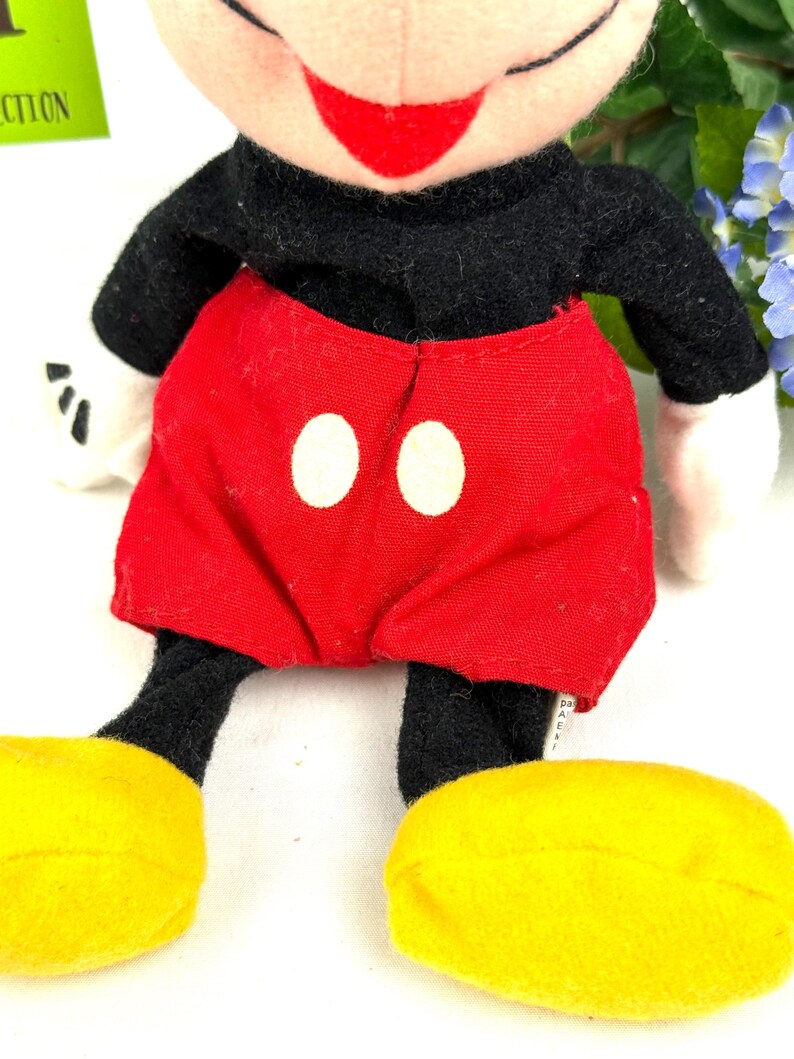 May include: A plush Mickey Mouse doll with a black body, red shorts with white buttons, yellow shoes, and white gloves. The doll has a smiling face with a red mouth and black eyes. The doll is a classic Disney character.