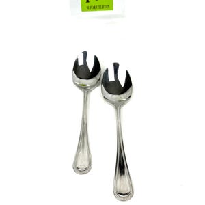 May include: Two shiny, silver salad spoons with a decorative beaded edge. The spoons have a unique design with a split end. A green sticker with the text "MY 90 YEAR COLLECTION" is in the background.