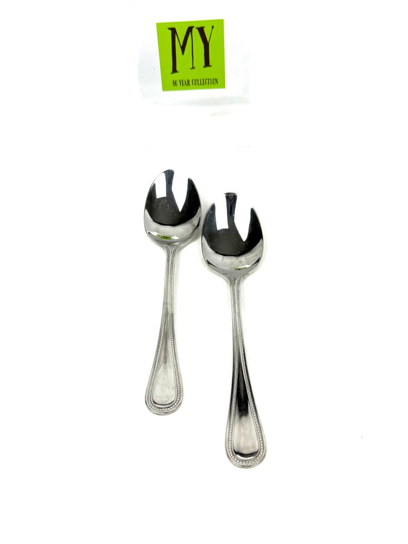 May include: Two shiny, silver salad spoons with a decorative beaded edge. The spoons have a unique design with a split end. A green sticker with the text "MY 90 YEAR COLLECTION" is in the background.