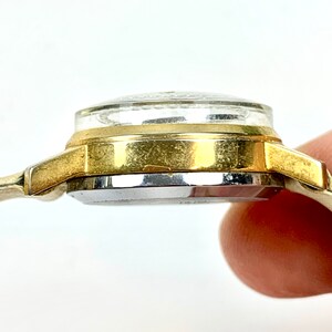 May include: Side view of a vintage gold-toned wristwatch. The watch features a clear, domed crystal, a gold-colored bezel, and a silver-colored watch face. The watch band is a thin, gold-toned metal.