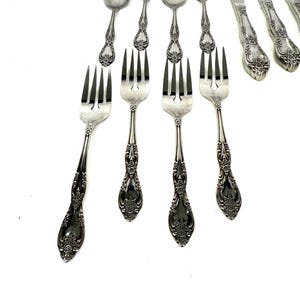 May include: A collection of ornate silver-colored flatware, including forks, spoons, and knives. The handles feature a detailed floral design. The utensils are arranged on a white surface, showcasing their elegant appearance.