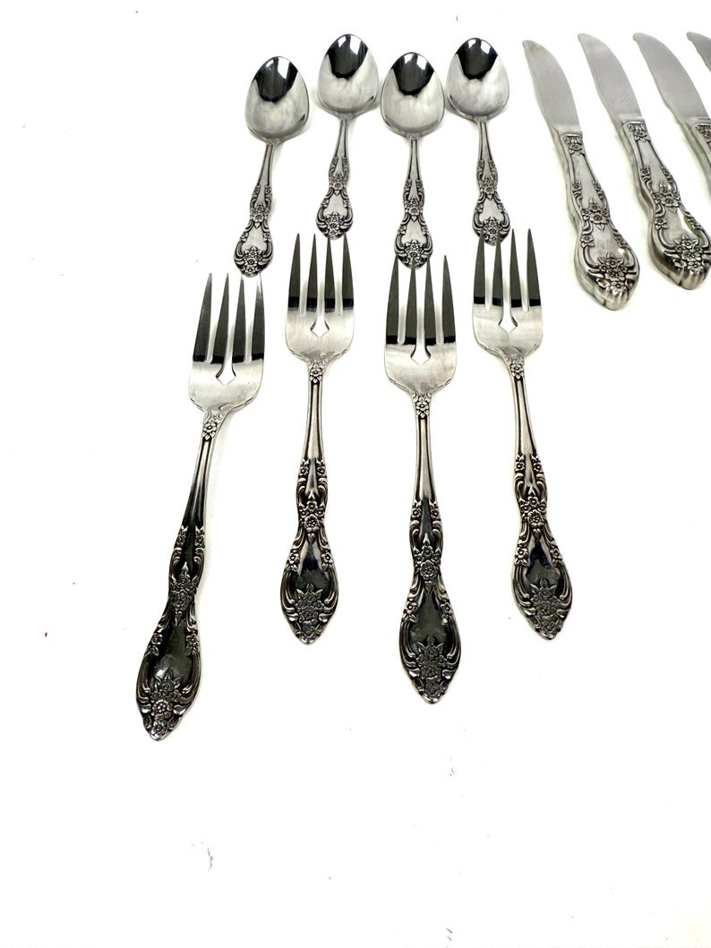 May include: A collection of ornate silver-colored flatware, including forks, spoons, and knives. The handles feature a detailed floral design. The utensils are arranged on a white surface, showcasing their elegant appearance.