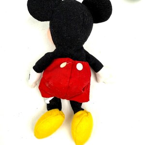 May include: A plush Mickey Mouse doll, viewed from the back. The doll features a black head with large ears, a red pair of shorts with white buttons, black arms and legs, white gloves, and yellow shoes. The doll is on a white background.