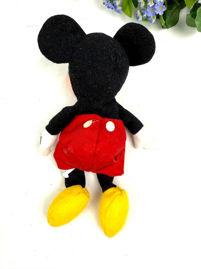 May include: A plush Mickey Mouse doll, viewed from the back. The doll features a black head with large ears, a red pair of shorts with white buttons, black arms and legs, white gloves, and yellow shoes. The doll is on a white background.