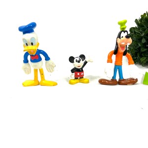 Może przedstawiać: Trzy wintażowe plastikowe figurki postaci Disneya: Donald Duck w niebieskim stroju marynarskim, Myszka Miki w czerwonych spodenkach i Goofy w pomarańczowej koszuli i niebieskich spodniach.