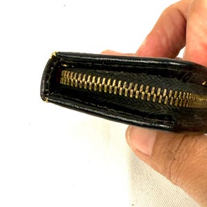 May include: Close-up of a black wallet with a gold zipper. The wallet appears to be made of leather or a similar material. The zipper is fully closed, and the wallet is slightly open, revealing a red interior.