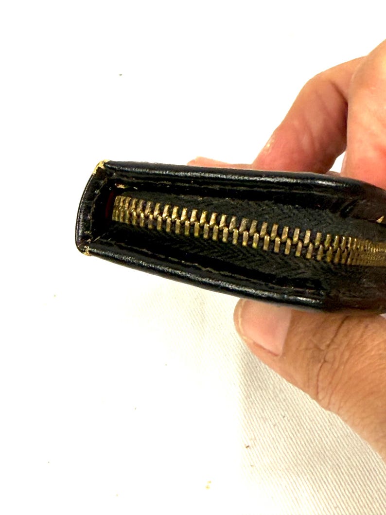 May include: Close-up of a black wallet with a gold zipper. The wallet appears to be made of leather or a similar material. The zipper is fully closed, and the wallet is slightly open, revealing a red interior.