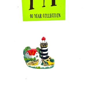 May include: A miniature lighthouse figurine with a white and black striped tower, a red roof, and a small house next to it. The figurine is set on a green base with yellow accents. The figurine is part of a 40 year collection.