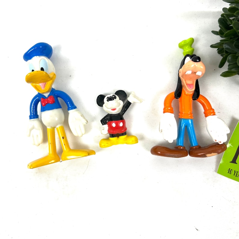 Może przedstawiać: Trzy plastikowe figurki postaci z Disneya: Donald Duck w niebieskim stroju marynarskim, Myszka Miki w czerwonej koszuli i czarnych spodenkach oraz Goofy w pomarańczowej koszuli i niebieskich spodniach.