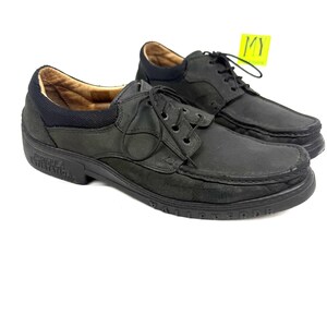 May include: A pair of black leather shoes with laces. The shoes have a rounded toe and a thick sole. The interior is a light brown color. A yellow tag is attached to one shoe. The shoes are displayed on a white background.