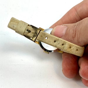 May include: A vintage gold-toned wristwatch with a cream-colored leather band. The watch face is silver with black hands and markers. The band shows signs of wear.