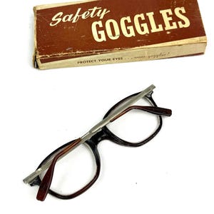 Pode incluir: &Oacute;culos de seguran&ccedil;a vintage com uma caixa de cart&atilde;o castanha. A caixa tem as palavras "Safety Goggles" em letras brancas. Os &oacute;culos t&ecirc;m arma&ccedil;&atilde;o preta, hastes castanhas e ponte prateada. O texto "Protect Your Eyes... wear goggles!" est&aacute; na caixa.