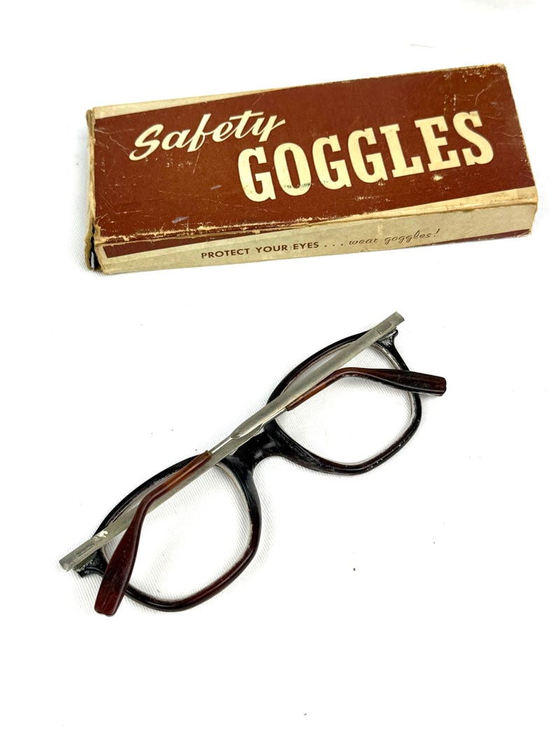 Pode incluir: &Oacute;culos de seguran&ccedil;a vintage com uma caixa de cart&atilde;o castanha. A caixa tem as palavras "Safety Goggles" em letras brancas. Os &oacute;culos t&ecirc;m arma&ccedil;&atilde;o preta, hastes castanhas e ponte prateada. O texto "Protect Your Eyes... wear goggles!" est&aacute; na caixa.