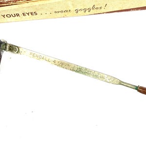 Pode incluir: Close-up de uma haste de &oacute;culos de seguran&ccedil;a vintage. A haste de metal est&aacute; marcada com "FENDALL 4 1/2". O texto "YOUR EYES... wear goggles!" &eacute; vis&iacute;vel em um fundo marrom.